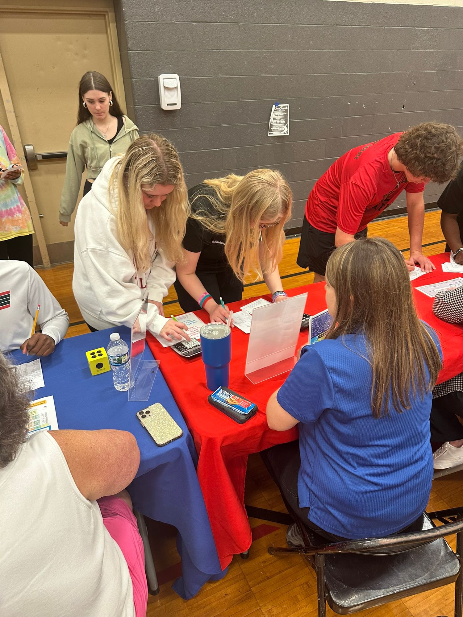 Students and volunteers gather around a red table in a school gymnasium, signing papers during a registration or sign-up event.