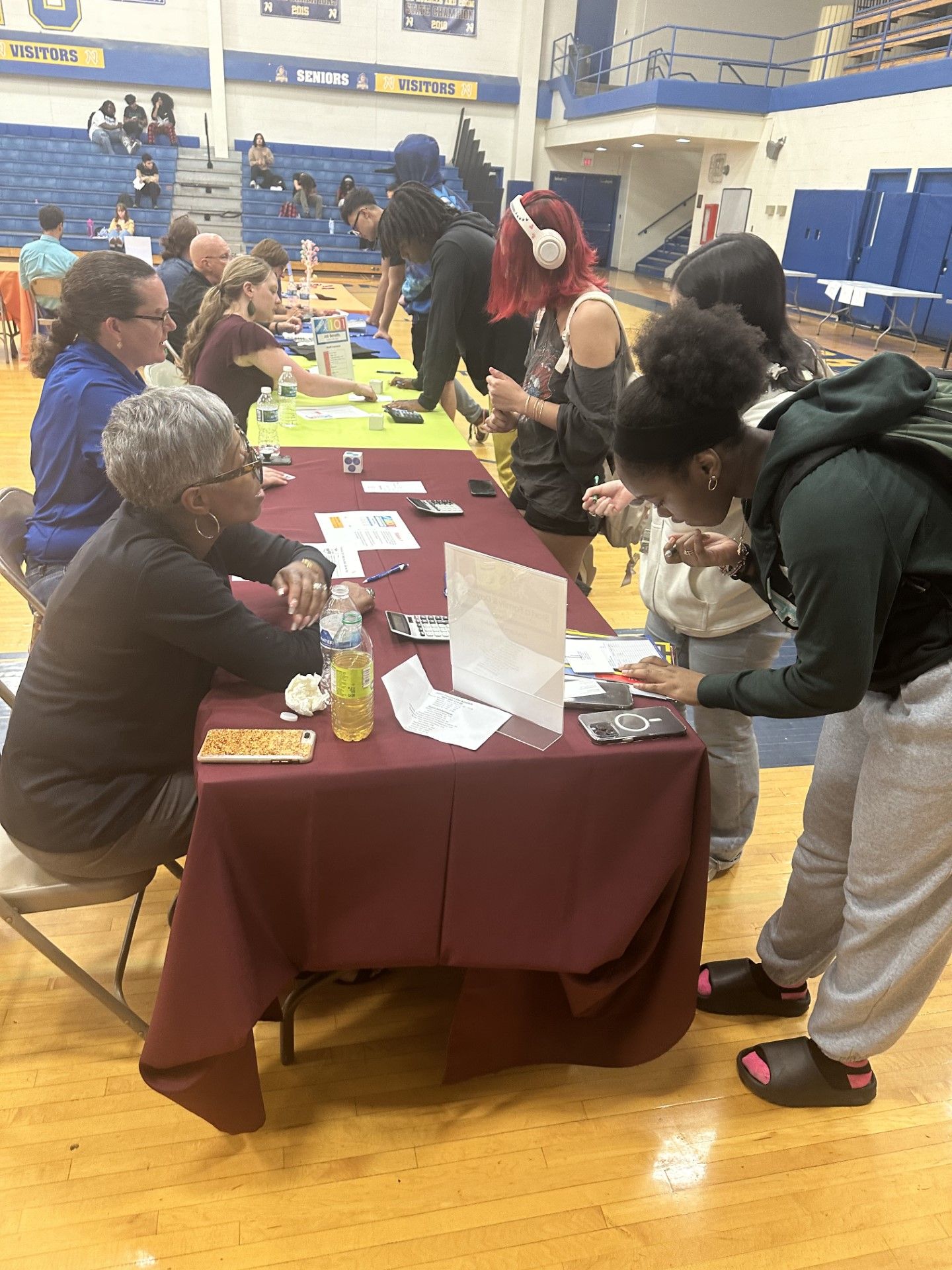 Students interact with advisors at a maroon-draped table during a high school gymnasium fair event.
