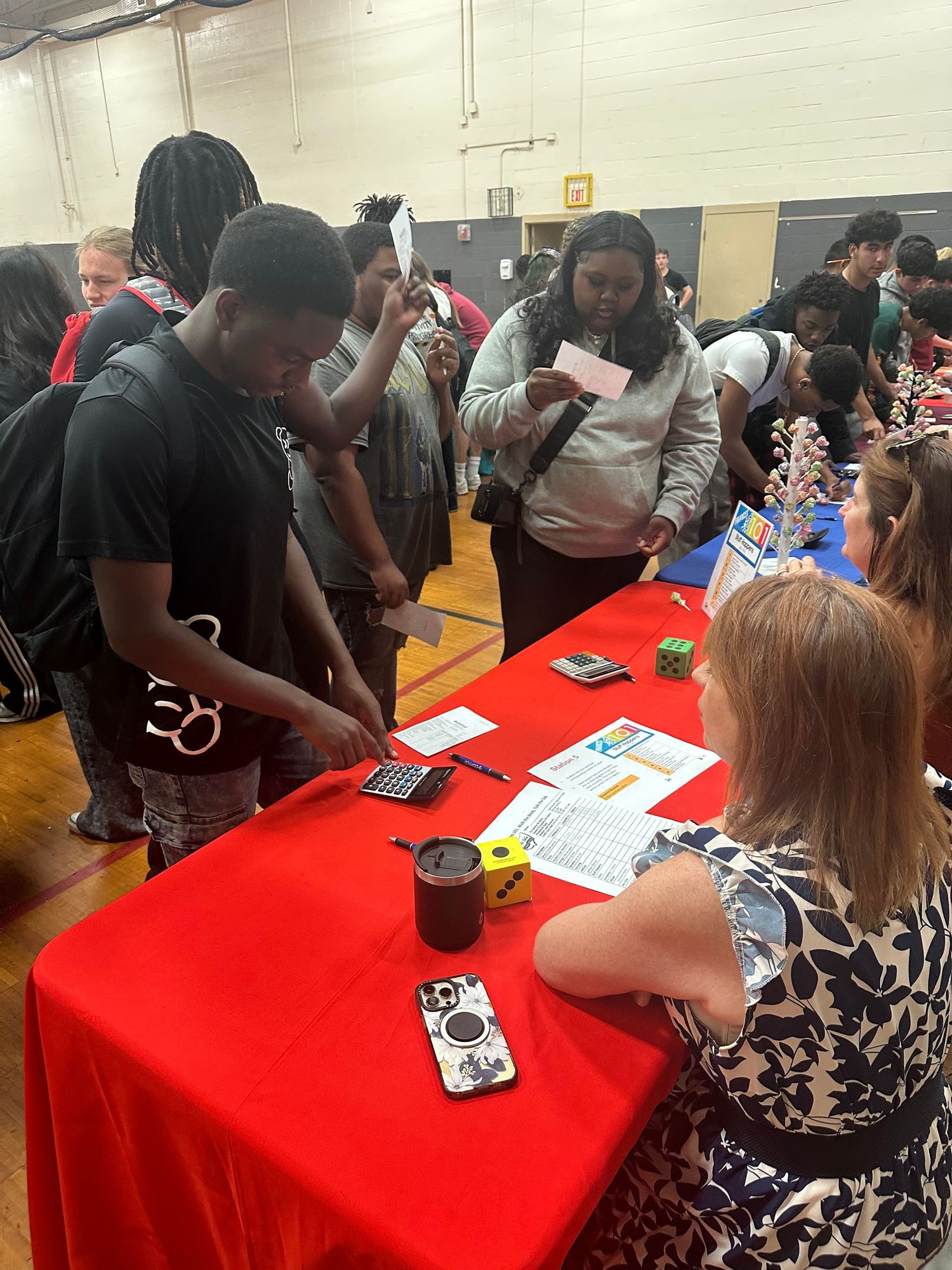 Students gather around a red table at a school career fair in a gymnasium, speaking with a recruiter.