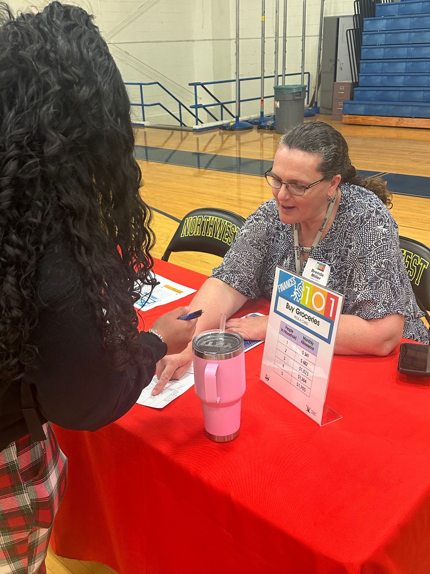 A woman with glasses and a name badge sits at a red table during a career fair in a school gymnasium.