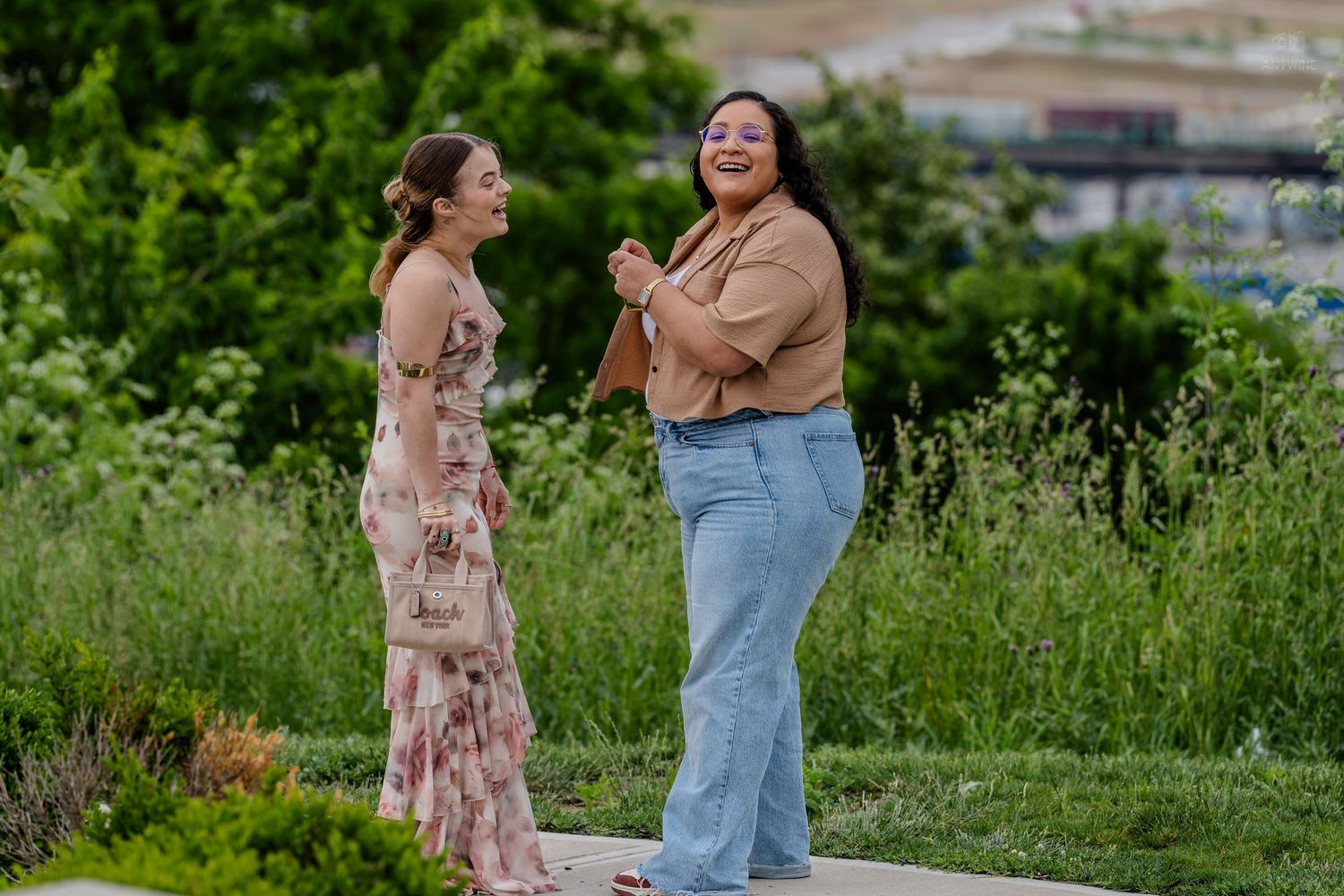 Two women laugh joyfully together outdoors in a lush green garden setting on a sunny day.