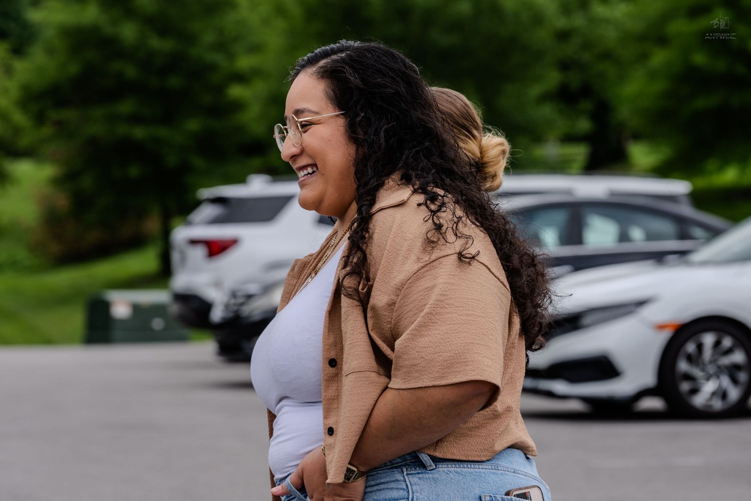 Young woman with curly hair and glasses laughing joyfully in an outdoor parking lot setting.