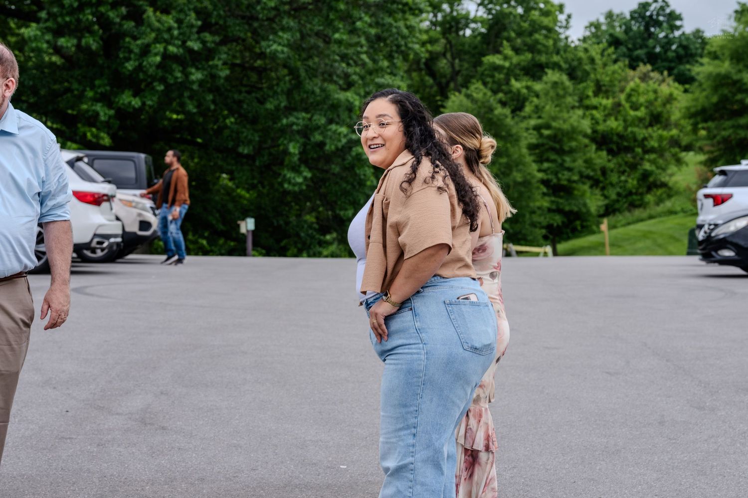 Smiling woman in jeans and tan top turning back in a parking lot with others nearby.