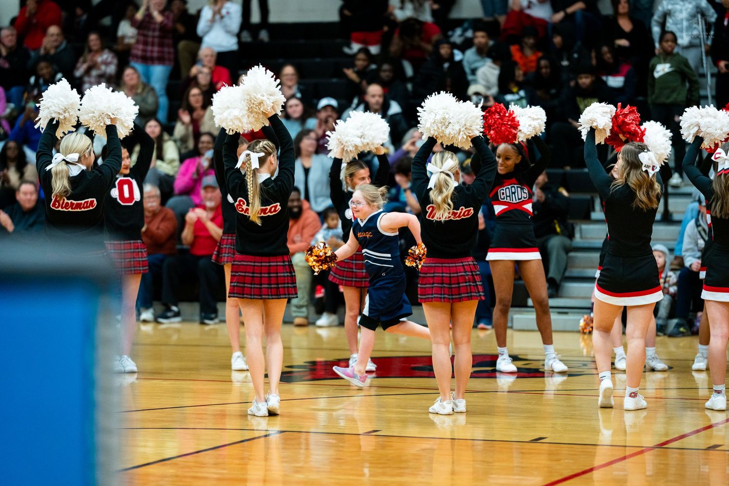 College cheerleaders perform with red and white pom-poms on a basketball court before an enthusiastic crowd.