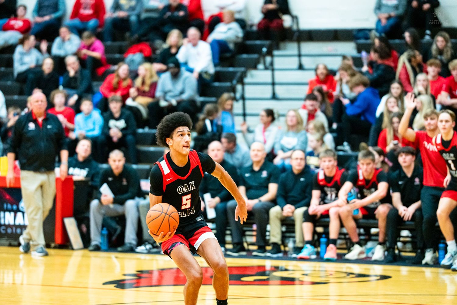 Young male basketball player wearing number 5 dribbles the ball during a high school game before a packed crowd.