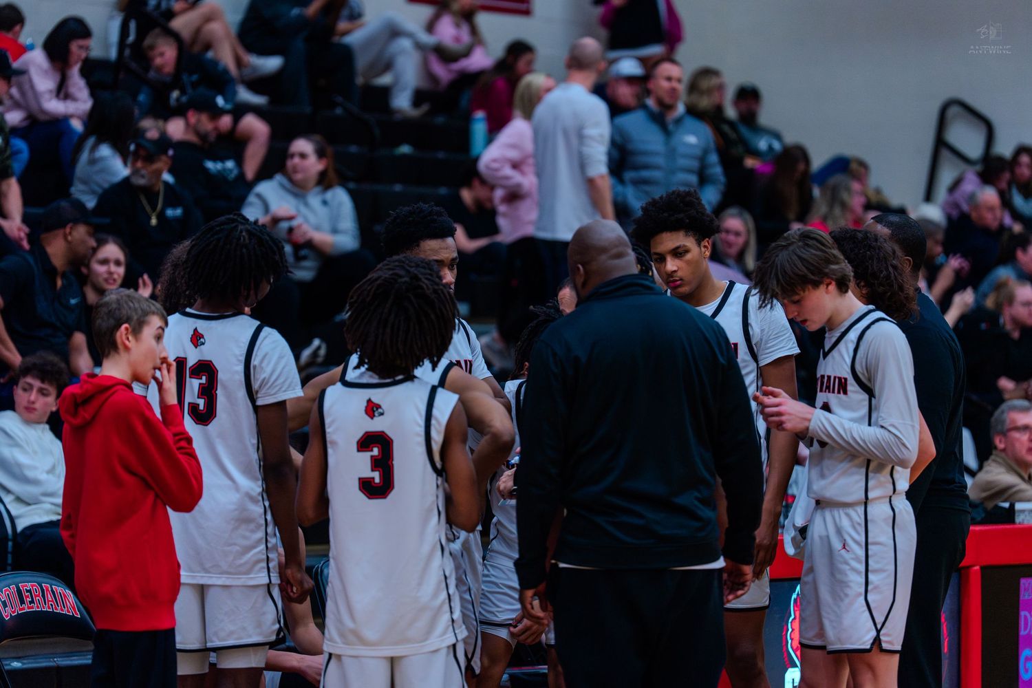 High school basketball team huddles with coach during timeout in packed gym.