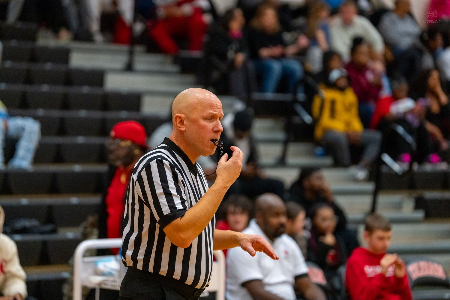 Bald basketball referee blows whistle and gestures during crowded gym game.