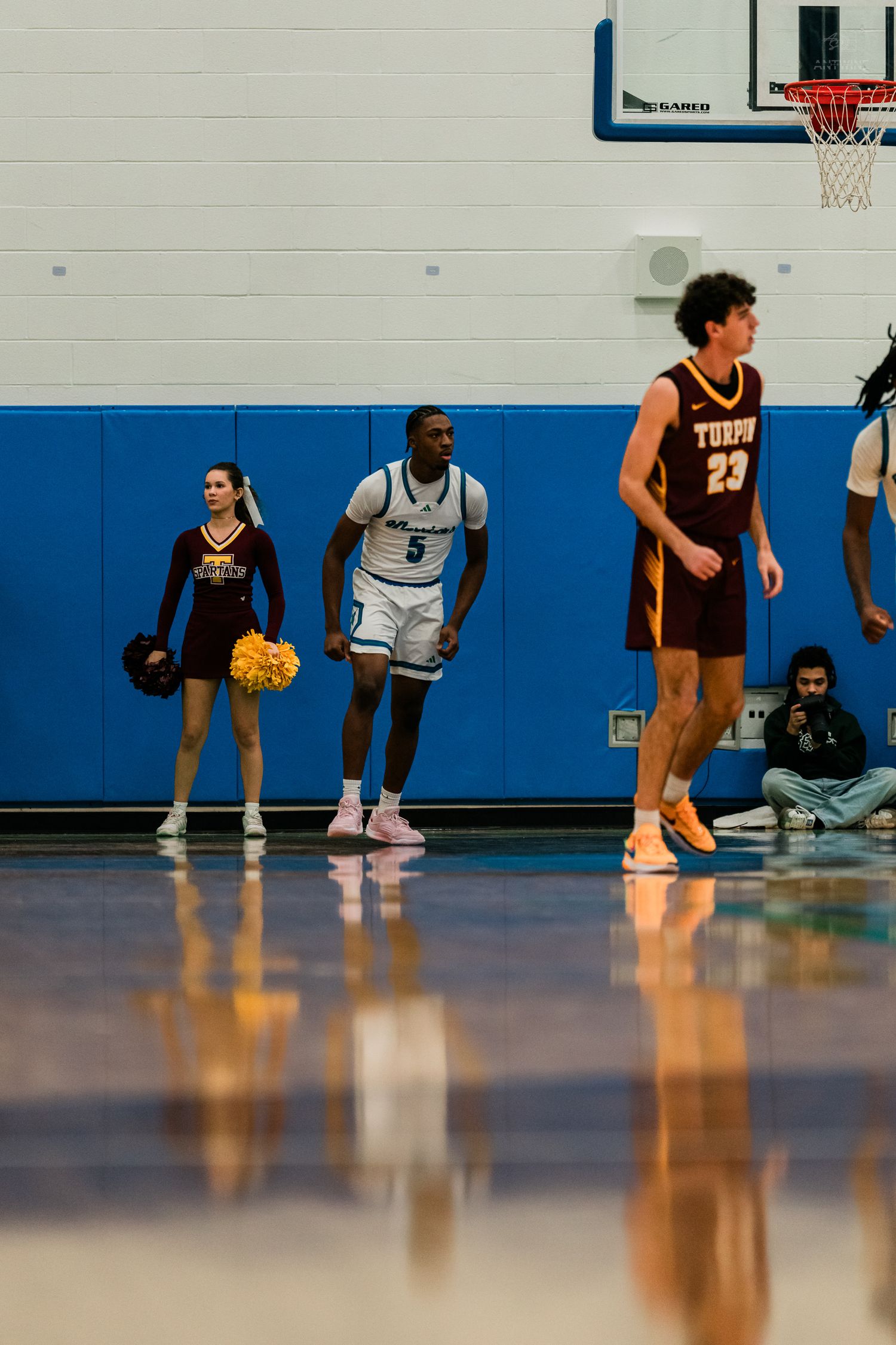 Turpin player number 23 stands near Warriors player 5 with cheerleader visible in blue gym background.