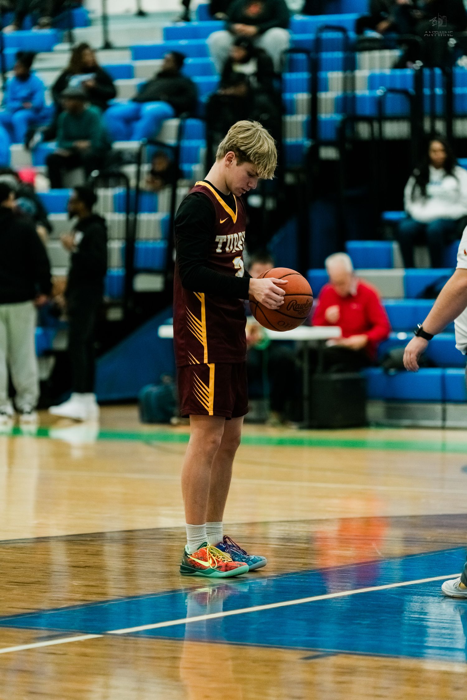 A blonde high school basketball player in a maroon uniform looks down at the ball on the court.