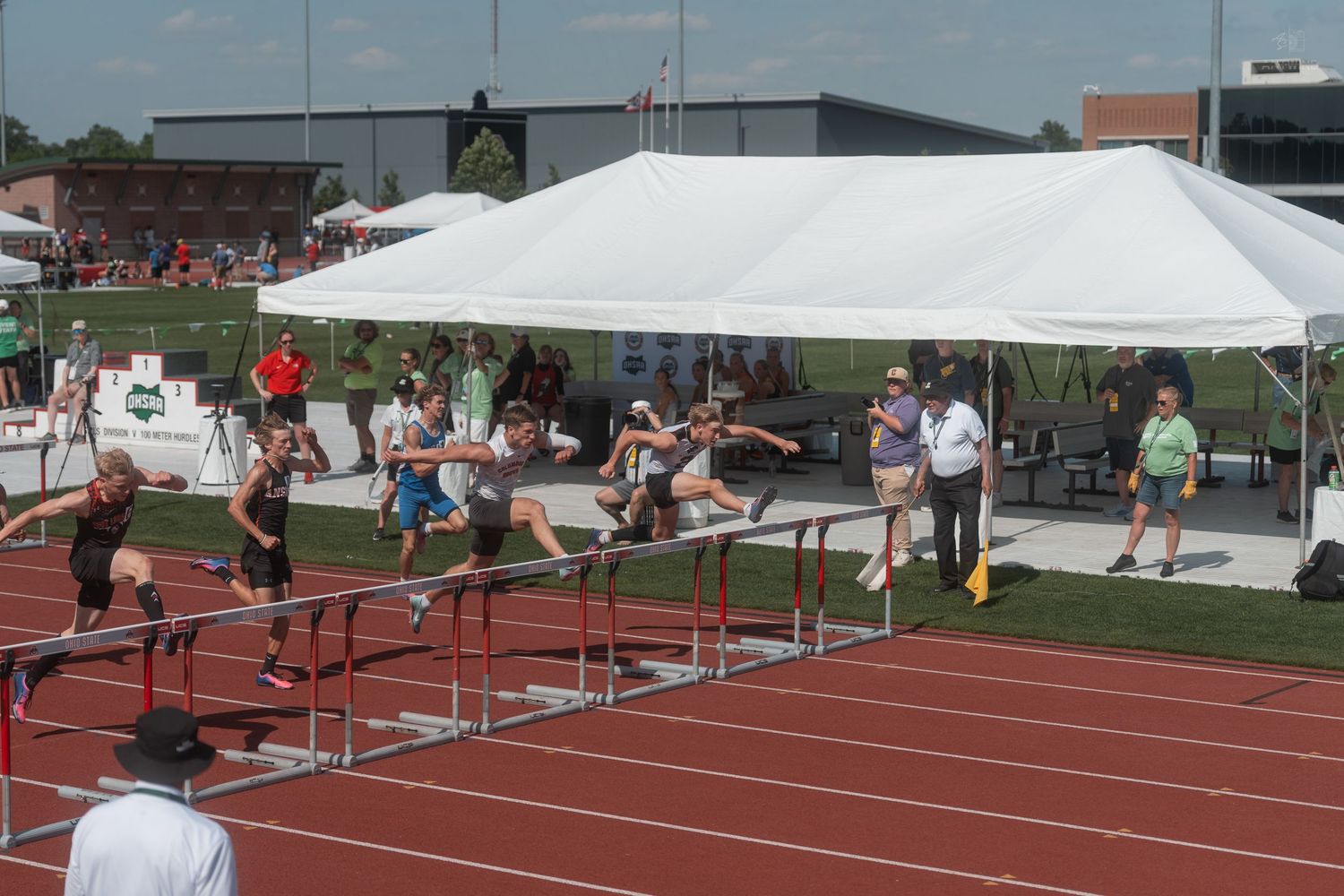 Young athletes leap over hurdles on a red track during an outdoor high school track and field competition.
