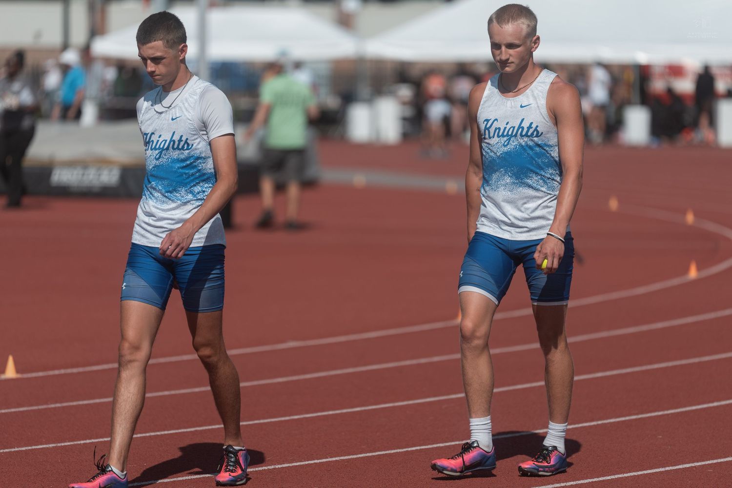 Two male athletes in blue Knights uniforms stand on a red track at a track meet.