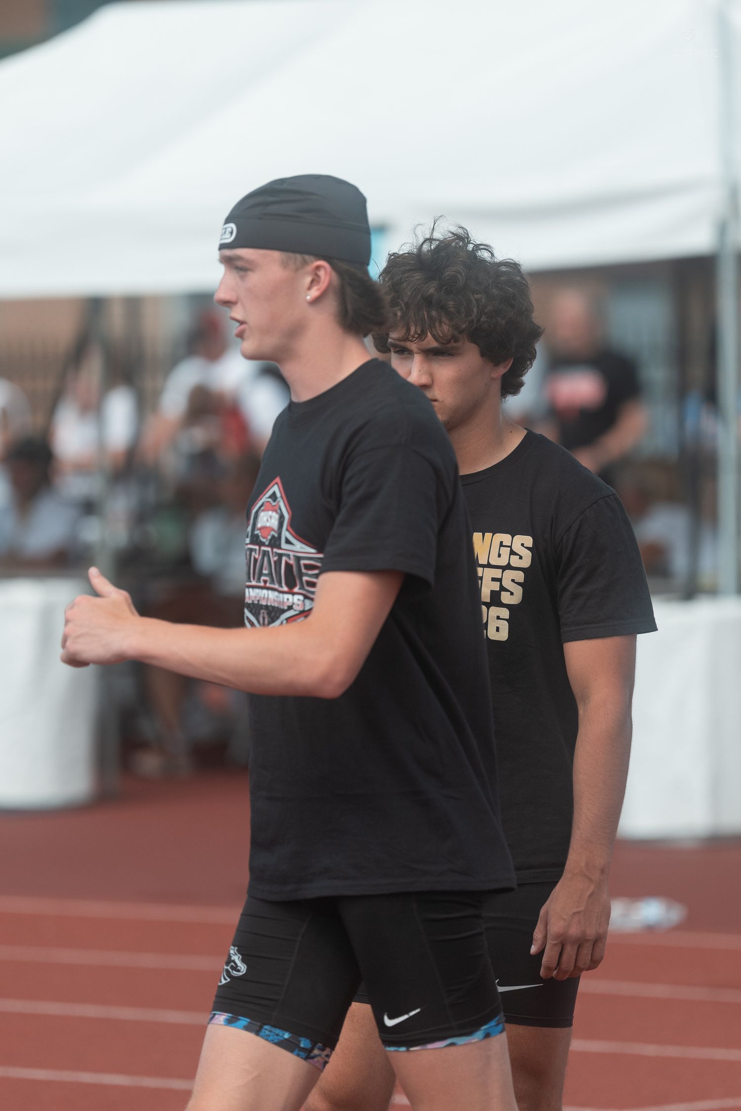 Two male athletes in black t-shirts stand trackside at an outdoor track and field competition event.