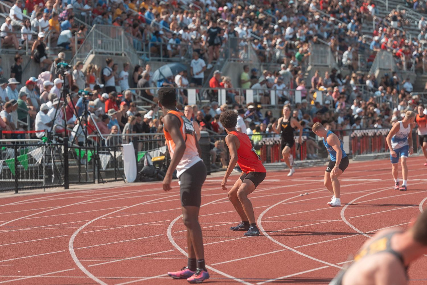 Track athletes exchange a baton during a relay race at a packed outdoor stadium track and field competition.