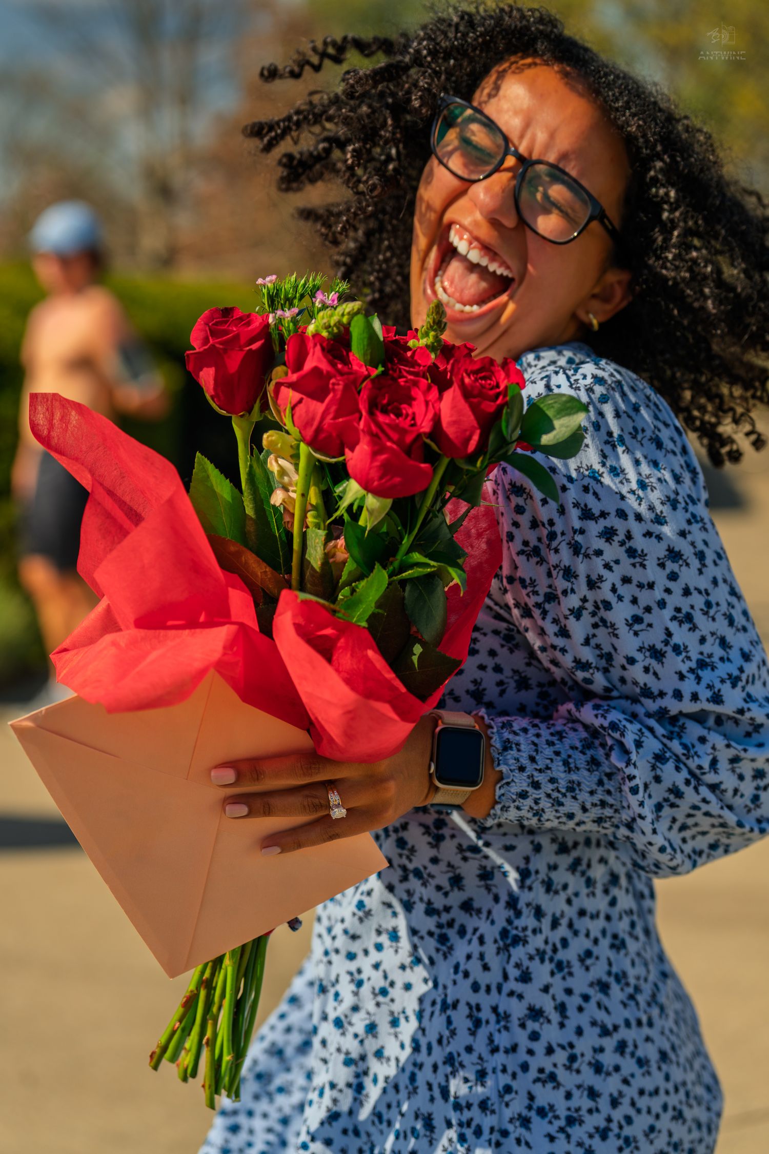 An ecstatic woman in a blue floral dress holds a bouquet of red roses while laughing outdoors.