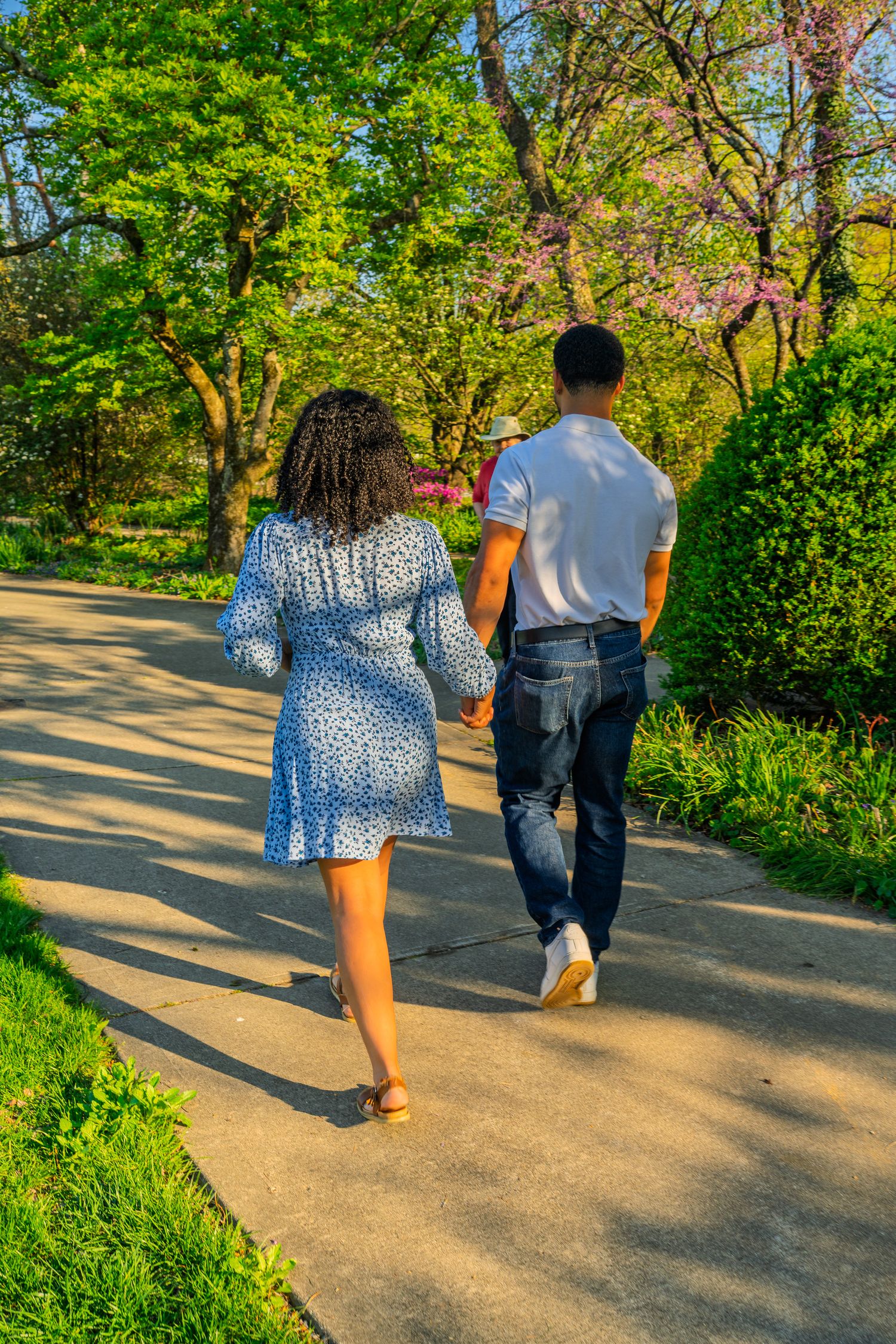 A couple holds hands while walking along a sunny park path surrounded by lush green trees and spring blossoms.