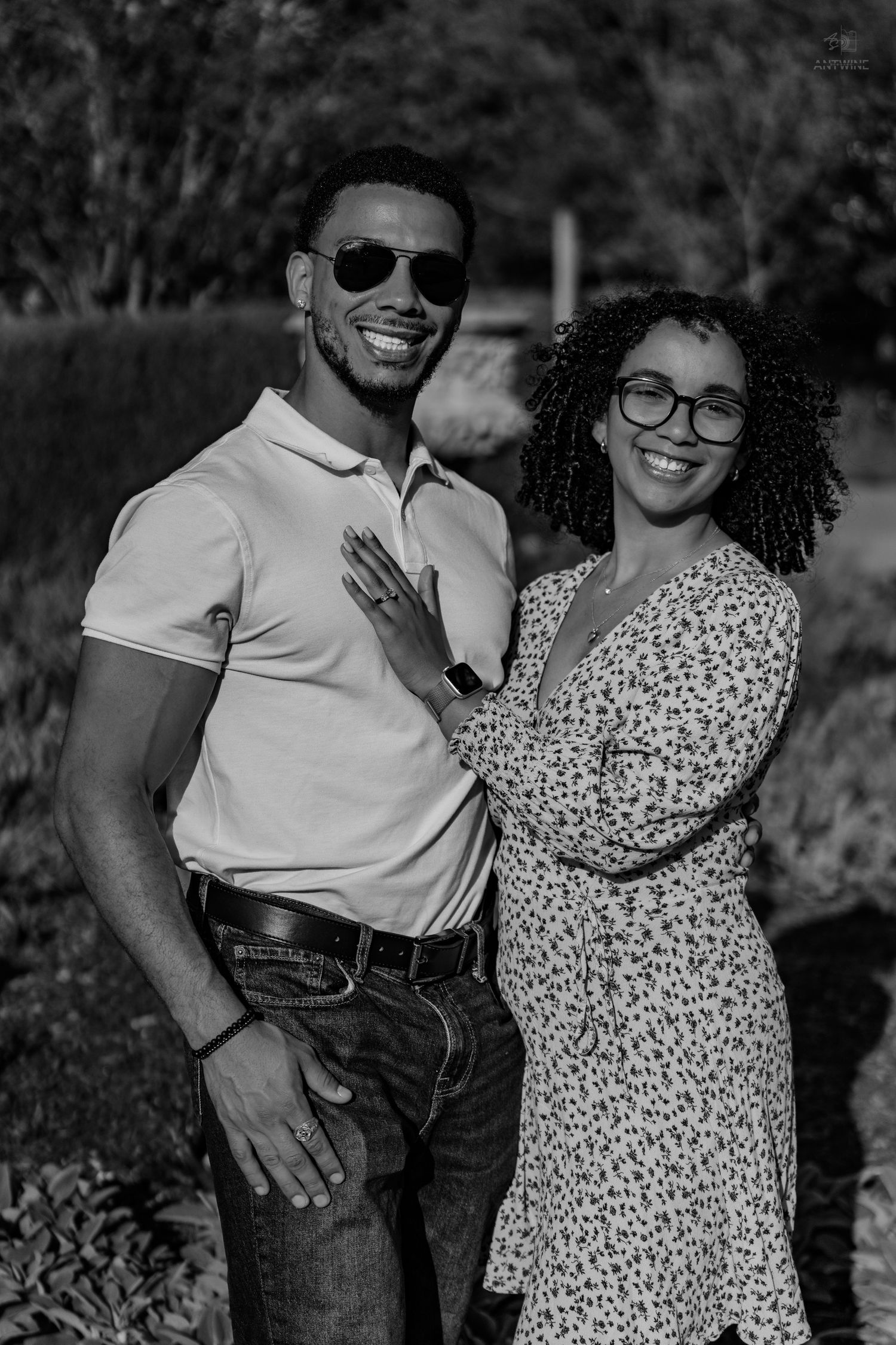 Black and white portrait of a smiling couple in a garden, man wearing sunglasses and woman in floral dress.