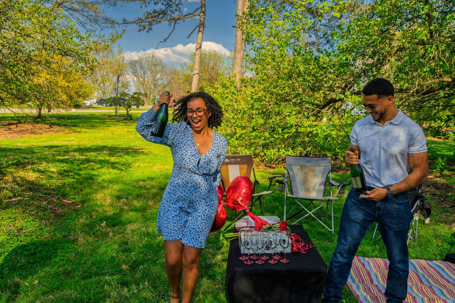 Joyful couple at outdoor picnic setup with red balloons, champagne glasses and decorative table in park.