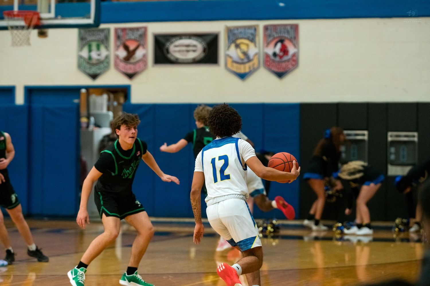 High school basketball player number 12 in white drives toward the basket while defended by green-uniformed opponent.