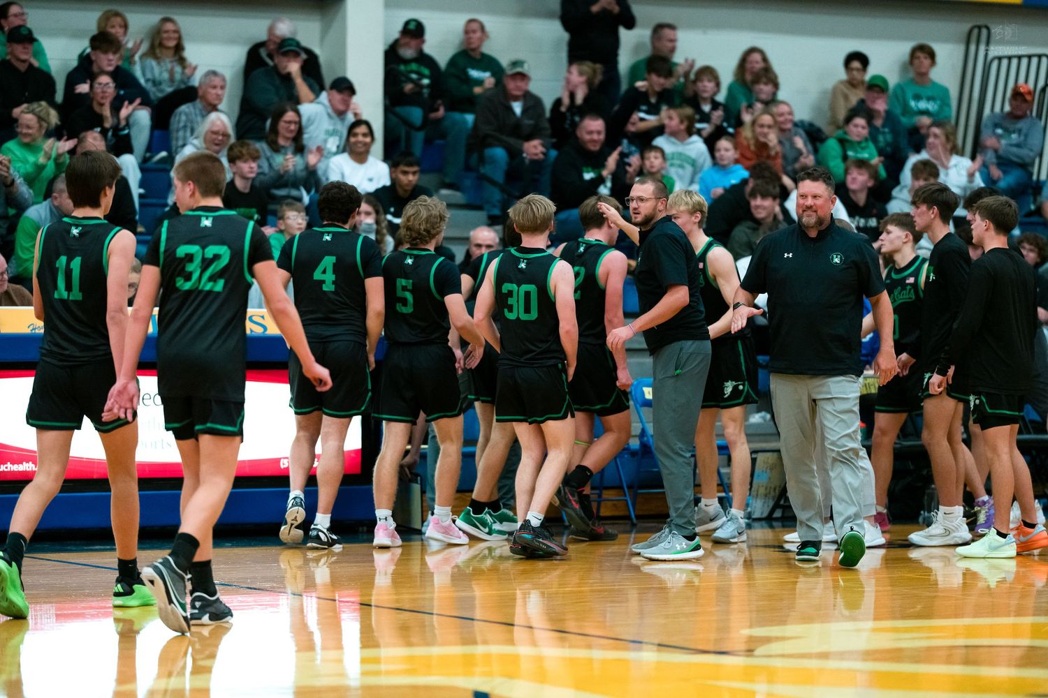 Green uniformed basketball team celebrates with coaches on court after winning gymnasium game.