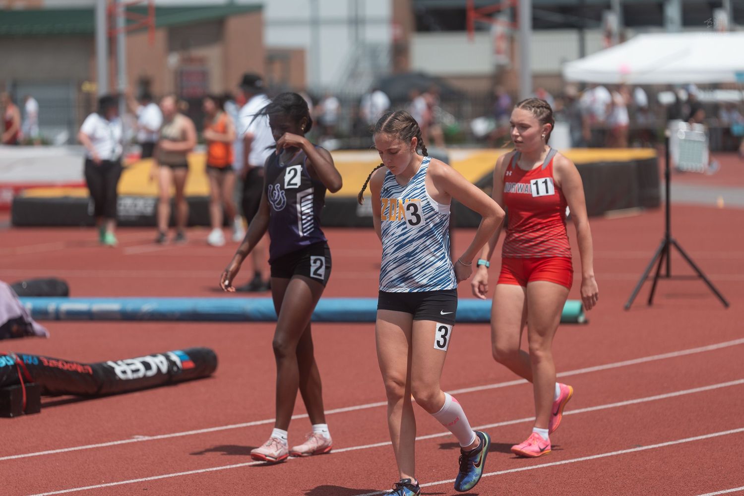 Female track athletes in numbered bibs walking on red track after completing a race at outdoor meet.