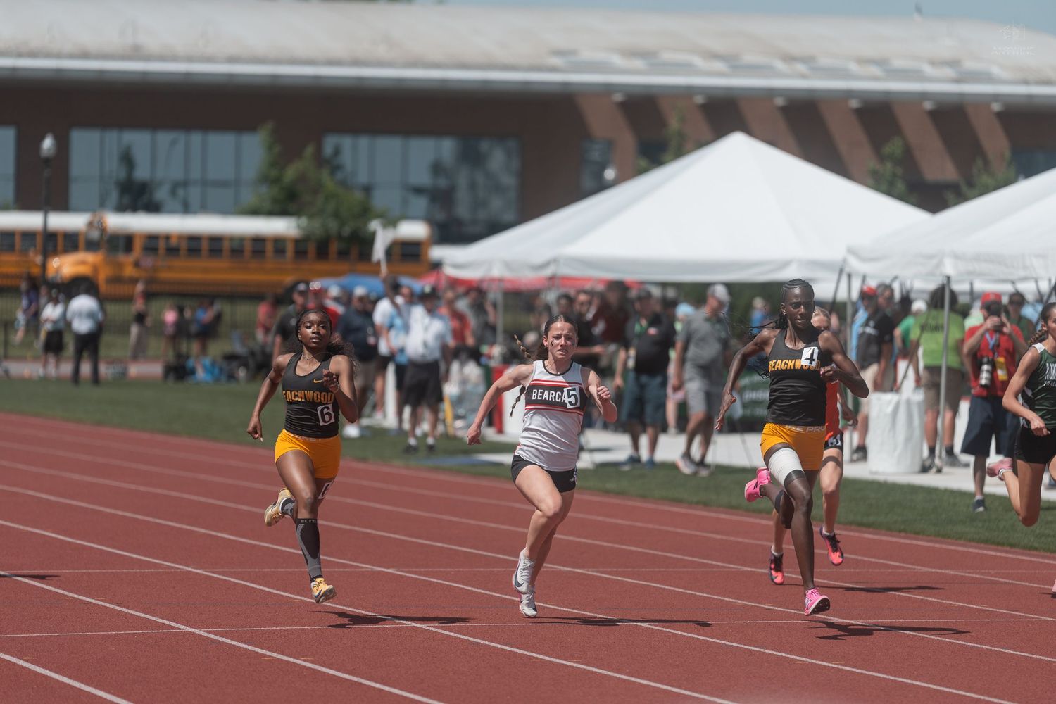 Female sprinters compete in a close race on a red track during a sunny outdoor track and field meet.