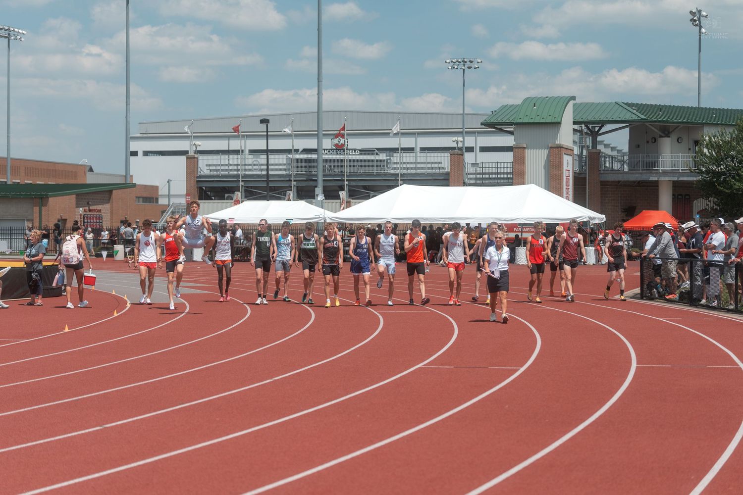 Track and field athletes line up at the starting line on a red track at an outdoor stadium.