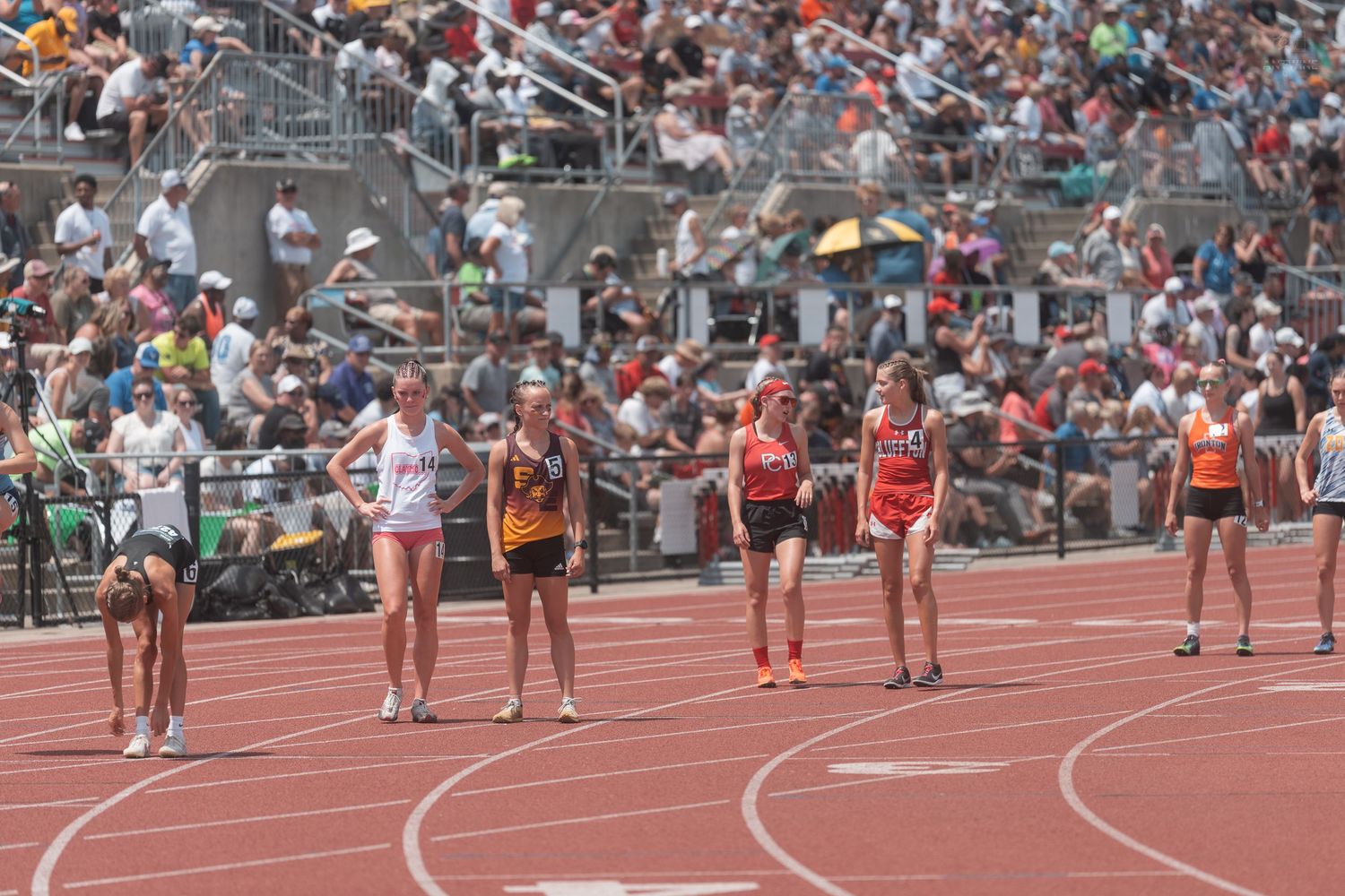 Female runners stand at the starting line on a red track during a sunny outdoor high school track competition.
