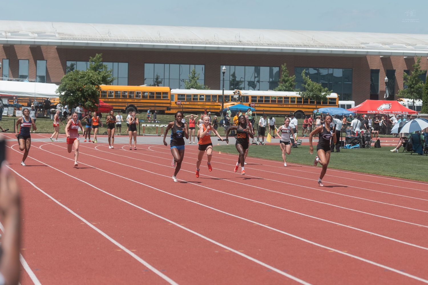 Athletes racing on red track at outdoor collegiate track and field meet with school buses in background.