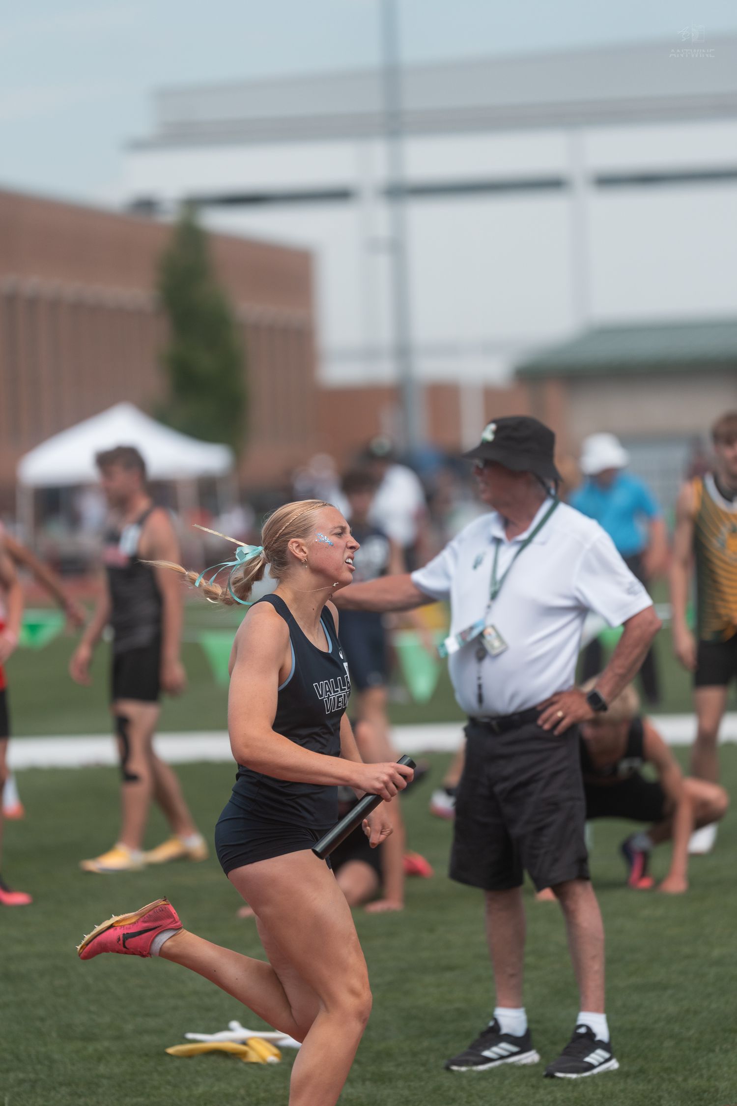 Young female sprinter in black uniform carries relay baton during outdoor high school track competition.