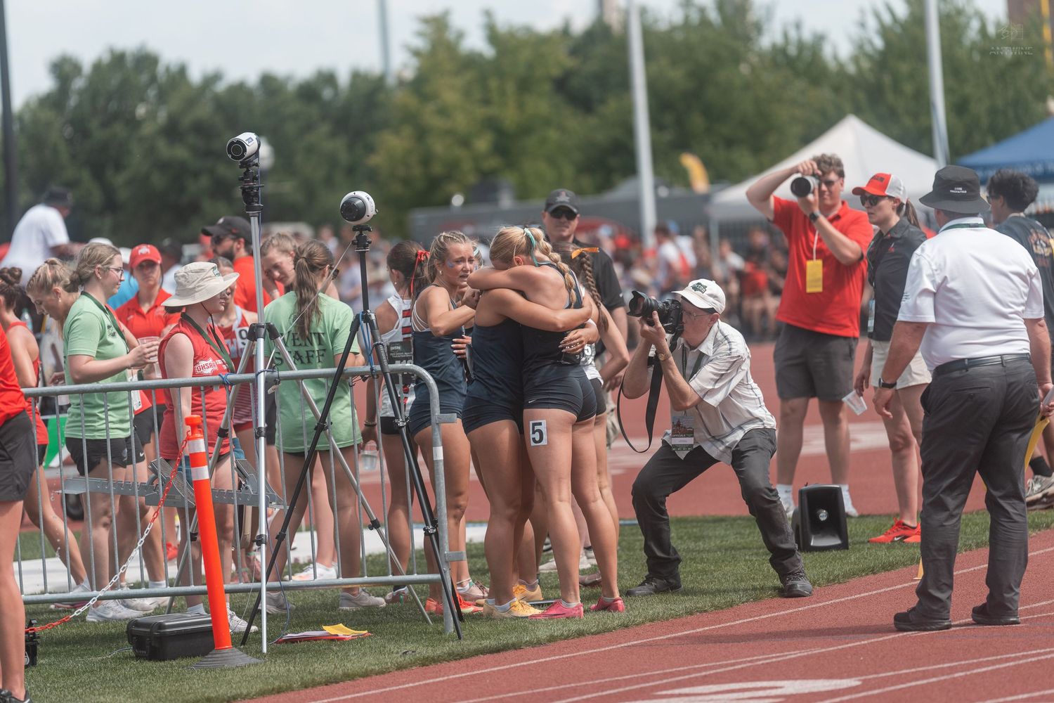 Girls track team embracing in celebration on the track while photographers capture the moment.
