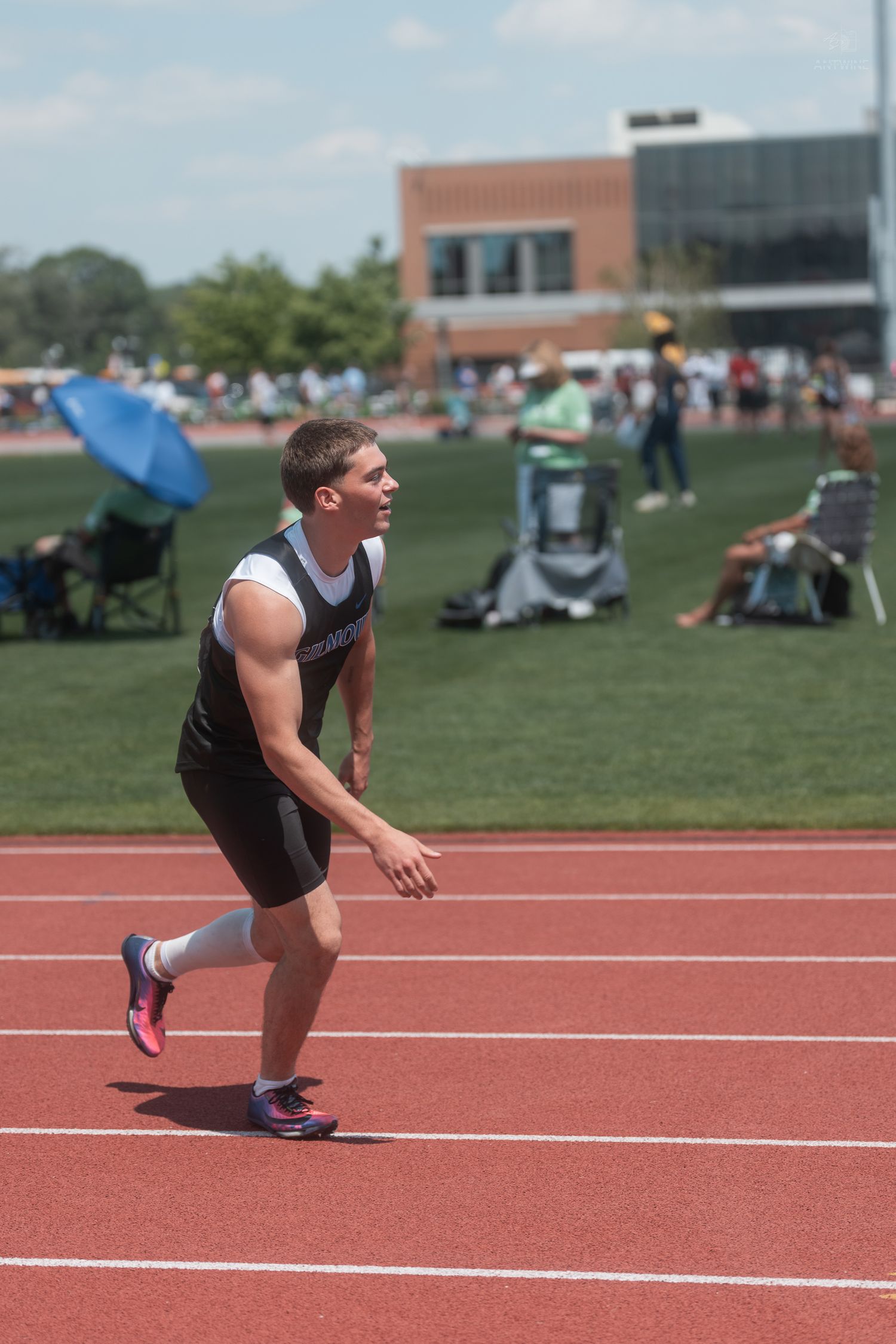 Male athlete in black uniform crouches in starting position on a red track at an outdoor track and field competition.