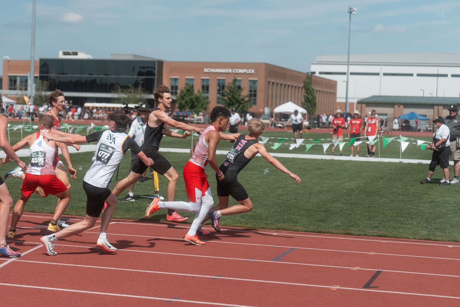 Athletes execute baton exchanges during relay race on green and red track at outdoor championship meet.