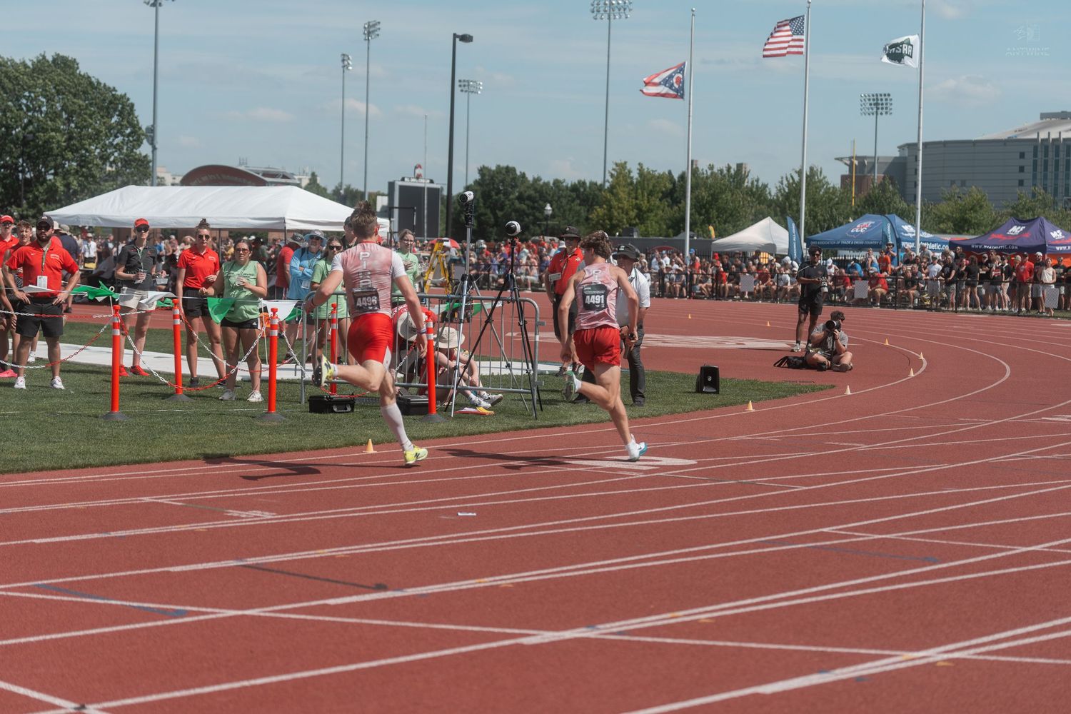Runners compete in relay race on red track with American flags and white tents visible in background.