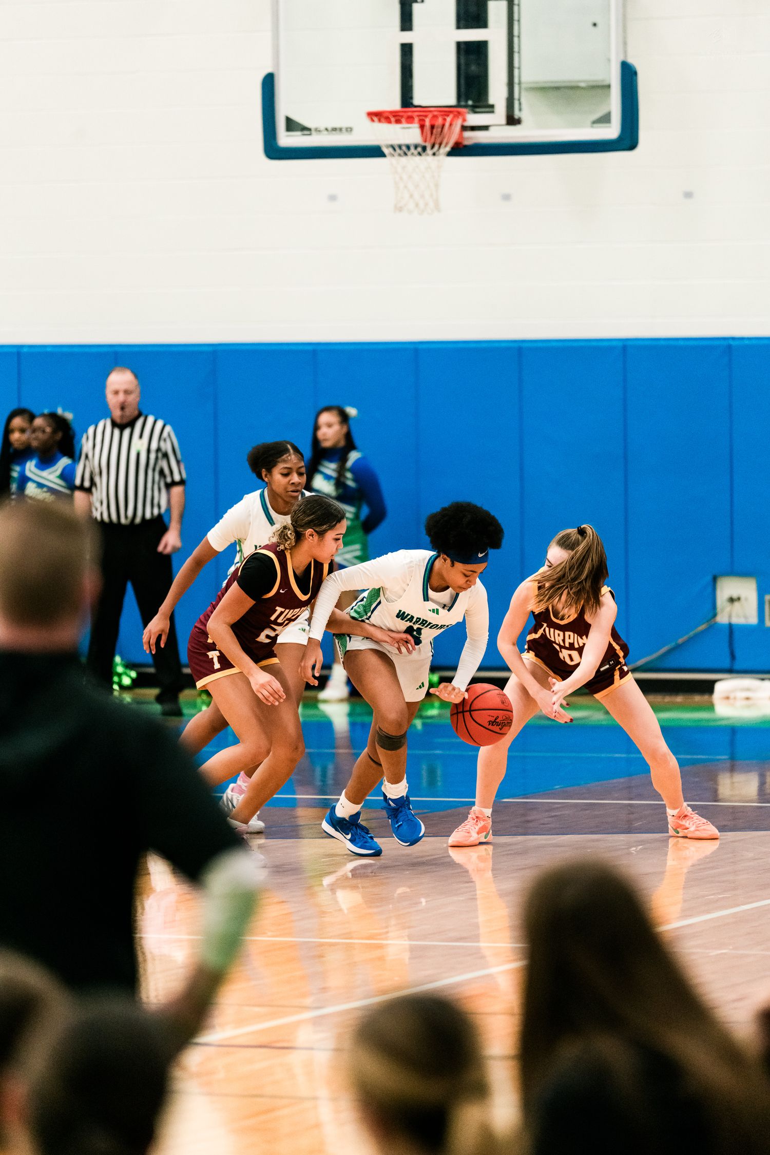 A Warriors player in white drives through two maroon-uniformed defenders during a girls high school basketball game.
