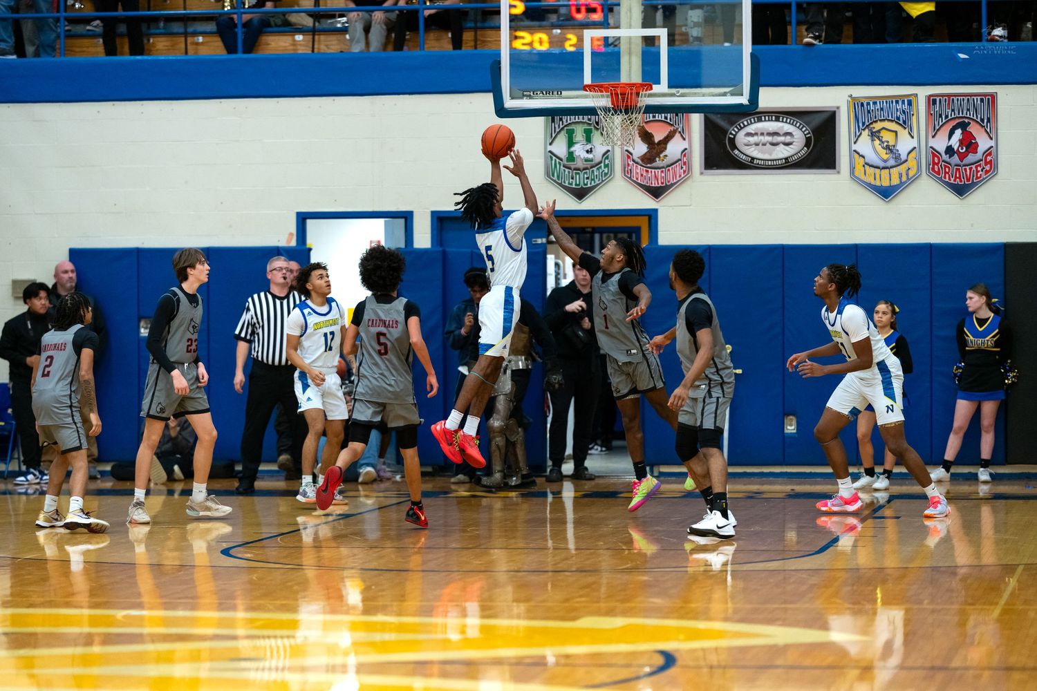 High school basketball player leaps for a shot near the basket during a game in a blue gymnasium.