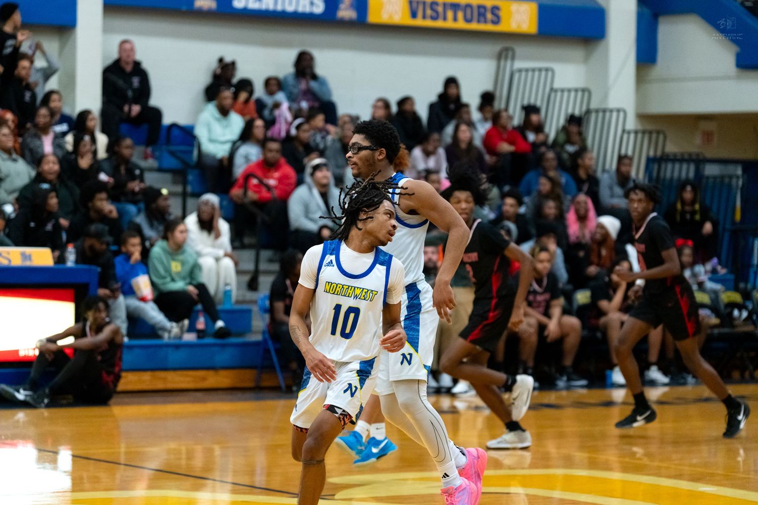 Two blue-uniformed basketball players #10 and teammate position near basket before packed home crowd.