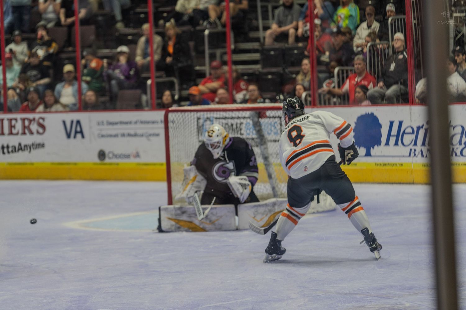Hockey player in orange jersey takes shot on goal against goalie in black pads during competitive ice hockey match.