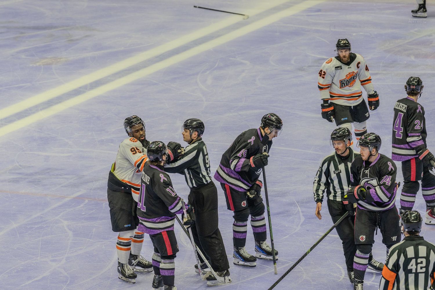 Ice hockey players from two teams gather near boards as referees intervene during game.