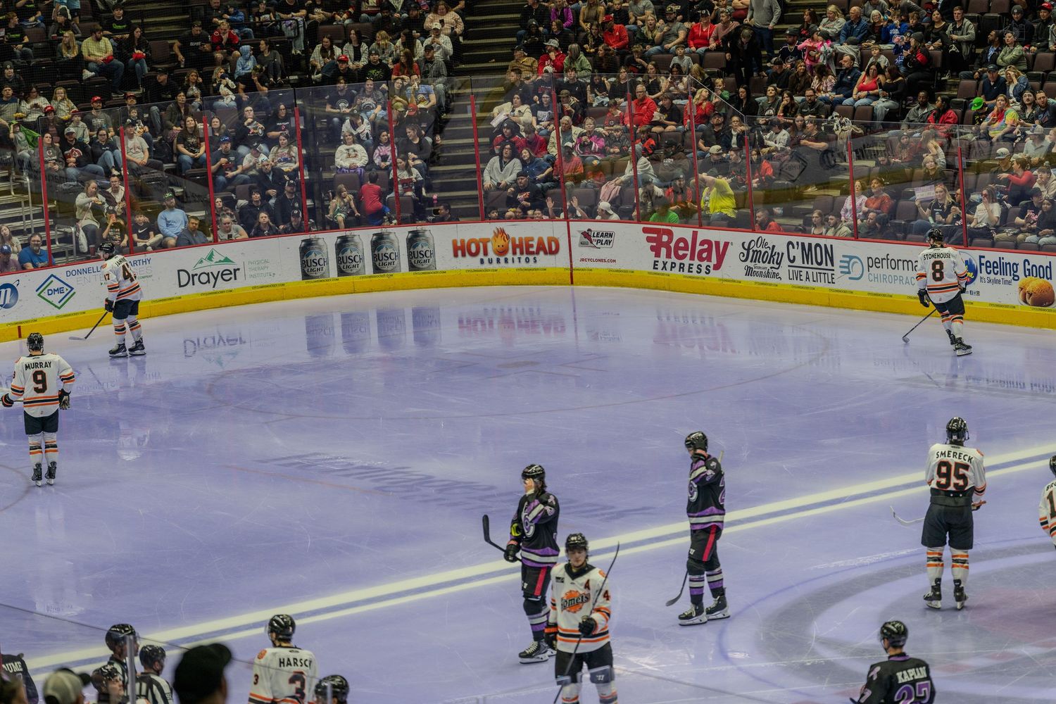 Hockey players from two teams stand on the ice during a stoppage in play at an indoor hockey arena.