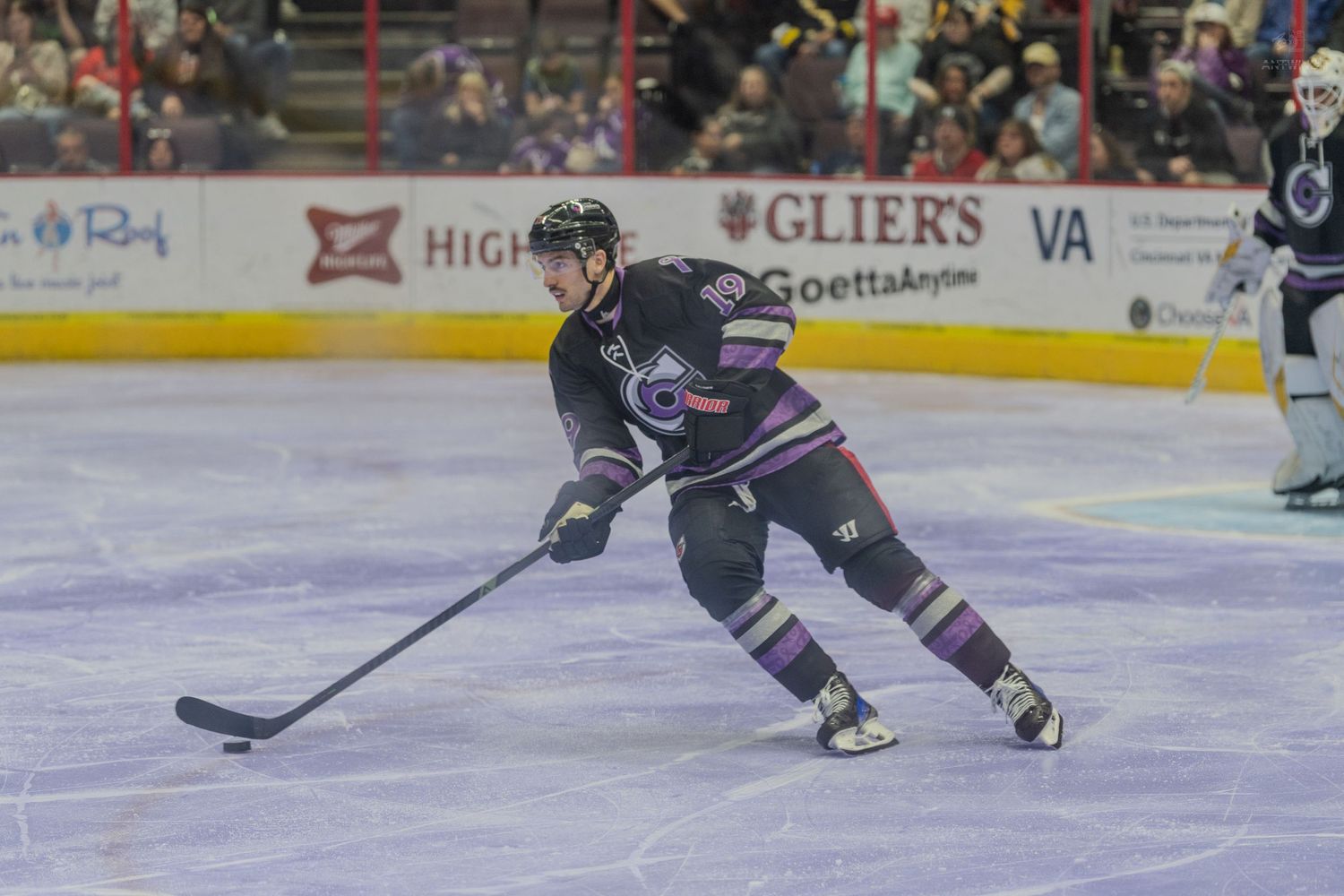 Hockey player in black and purple uniform skating with the puck during a professional hockey game.
