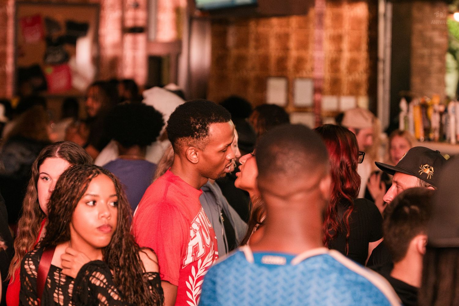 Two people share an intimate moment in a busy nightclub crowd with colorful ambient lighting.