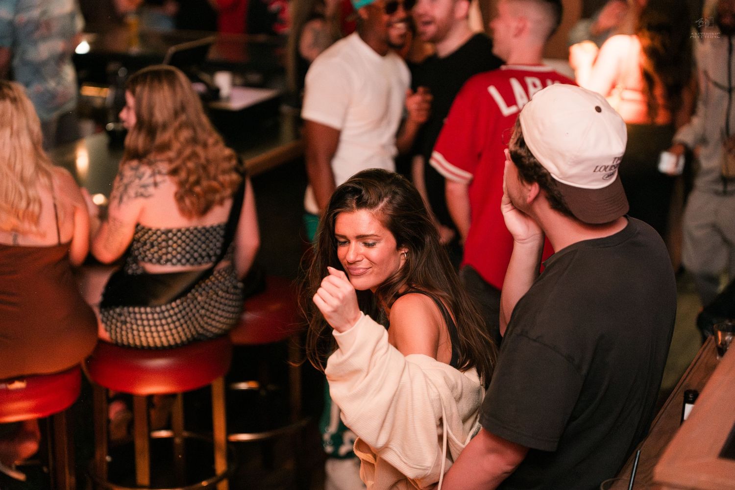 Young woman in cream hoodie laughing with a man in a white cap at a bar.