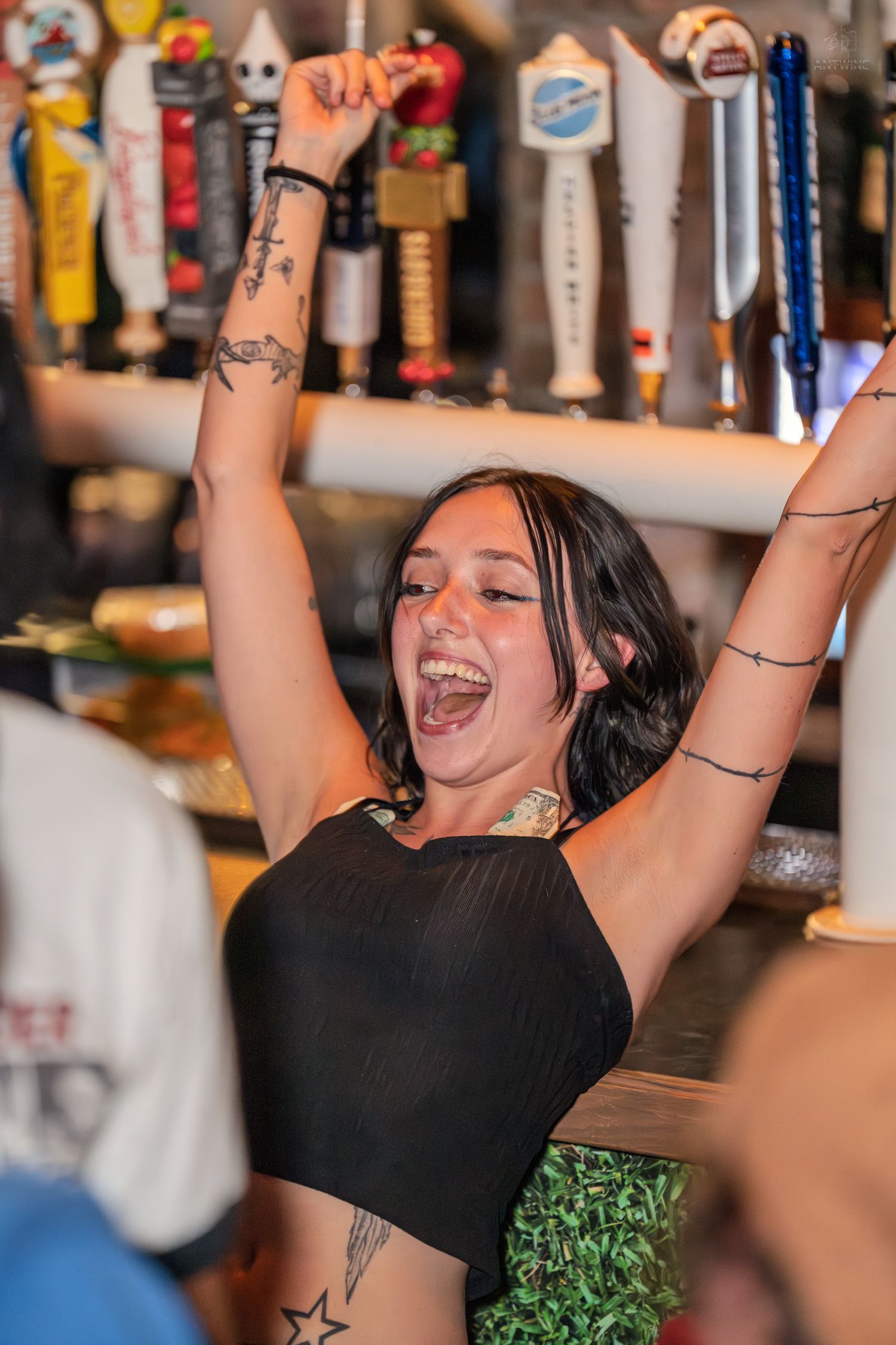 Excited young tattooed woman celebrating with arms raised near beer taps at a lively bar.