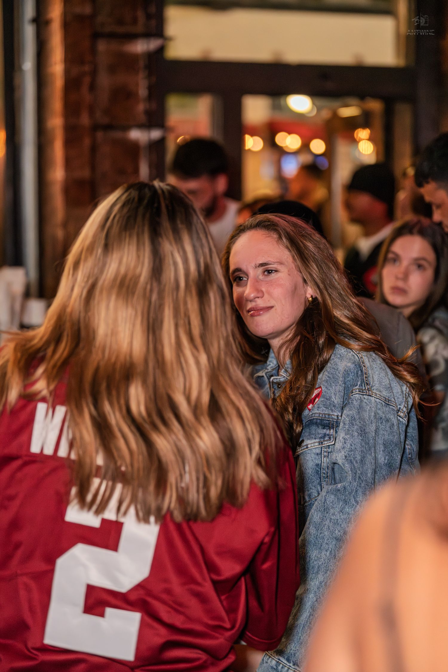 Two young women chatting at an outdoor bar wearing a denim jacket and sports jersey.