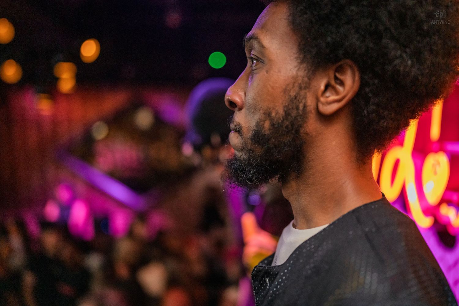 A bearded Black man in profile view at a vibrant nightclub with colorful neon lights in the background.