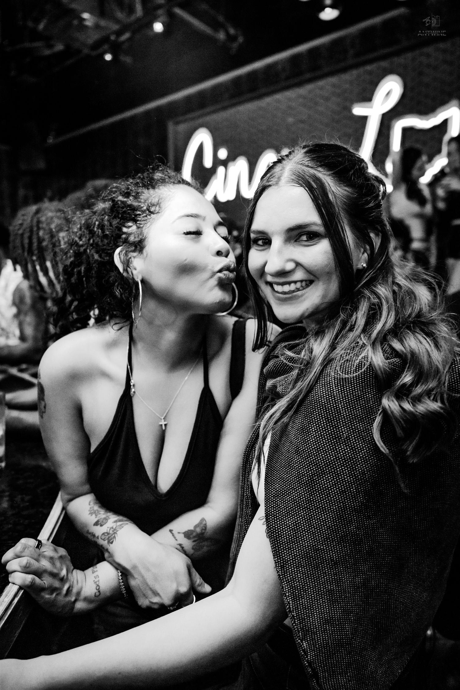 Two women posing together at a bar with neon signage in black and white photo.