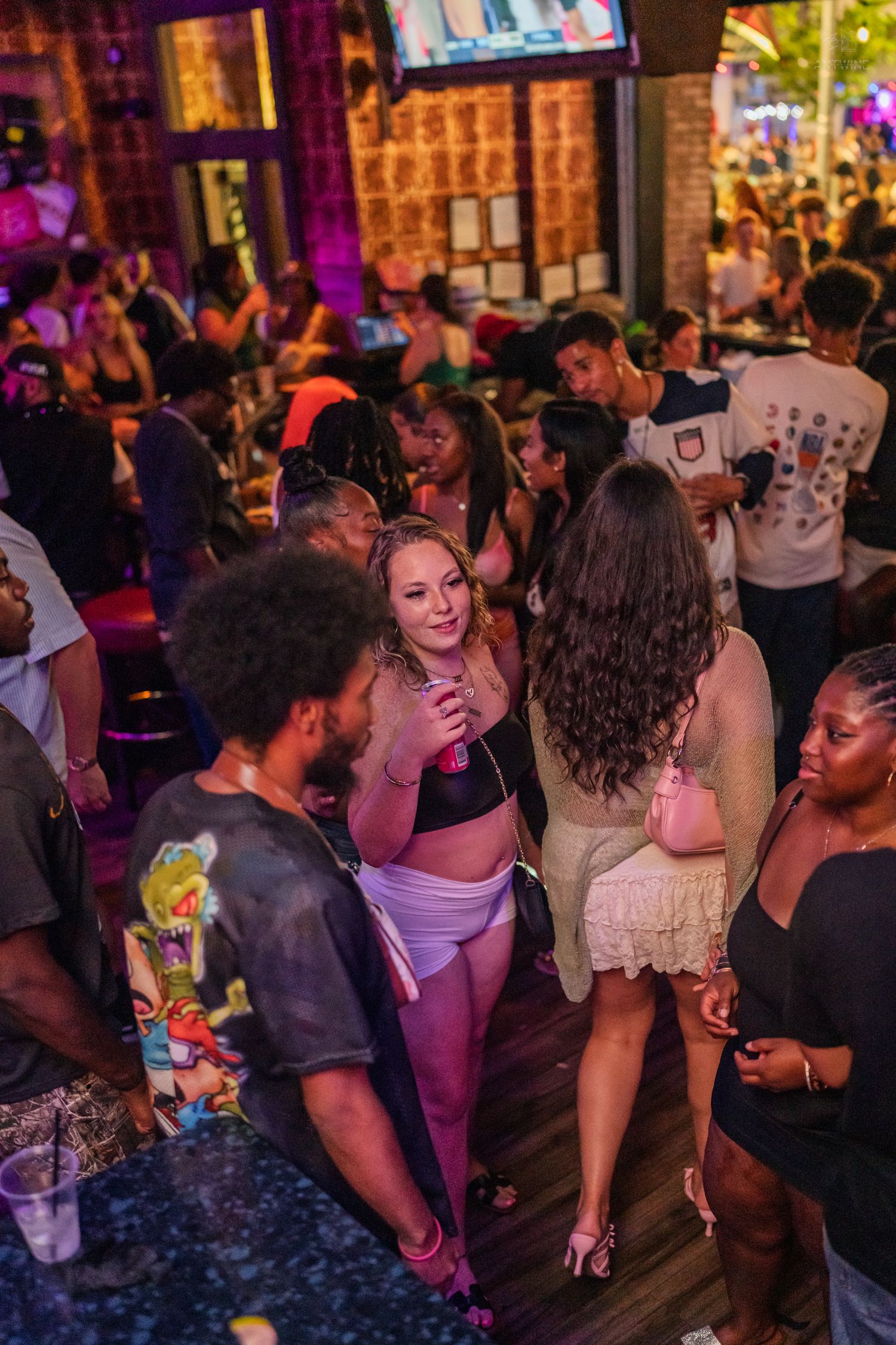 Diverse group of partygoers socializing under purple lights at a packed nightclub bar.