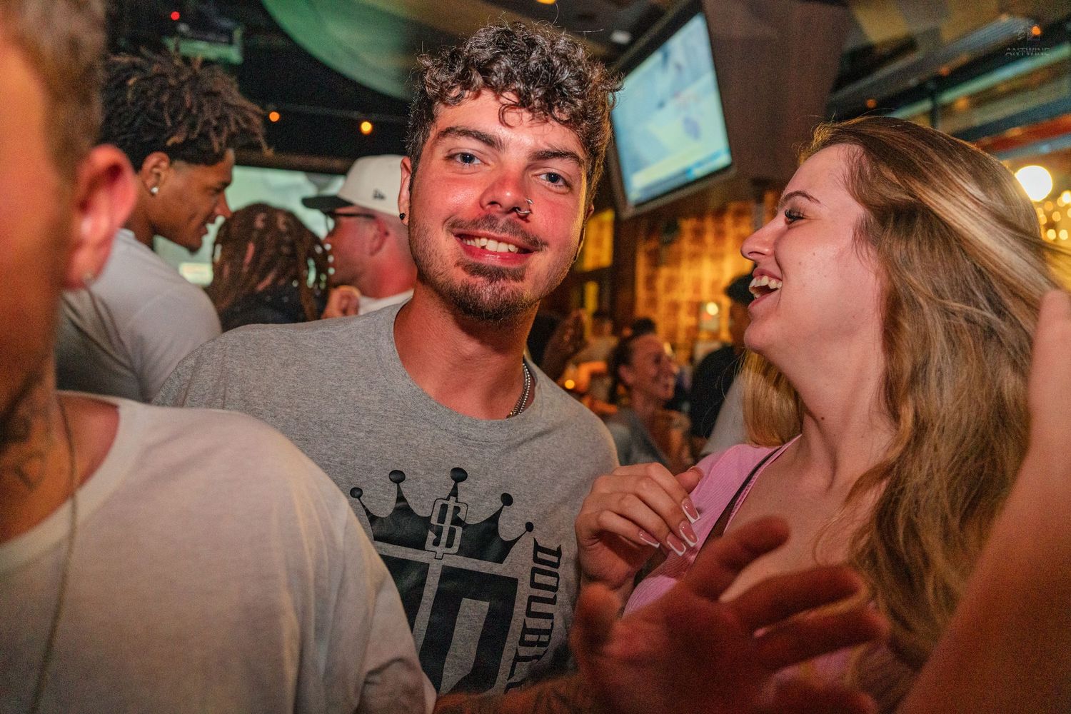 Young man in gray graphic tee smiling at a lively bar with friends and TVs in background.