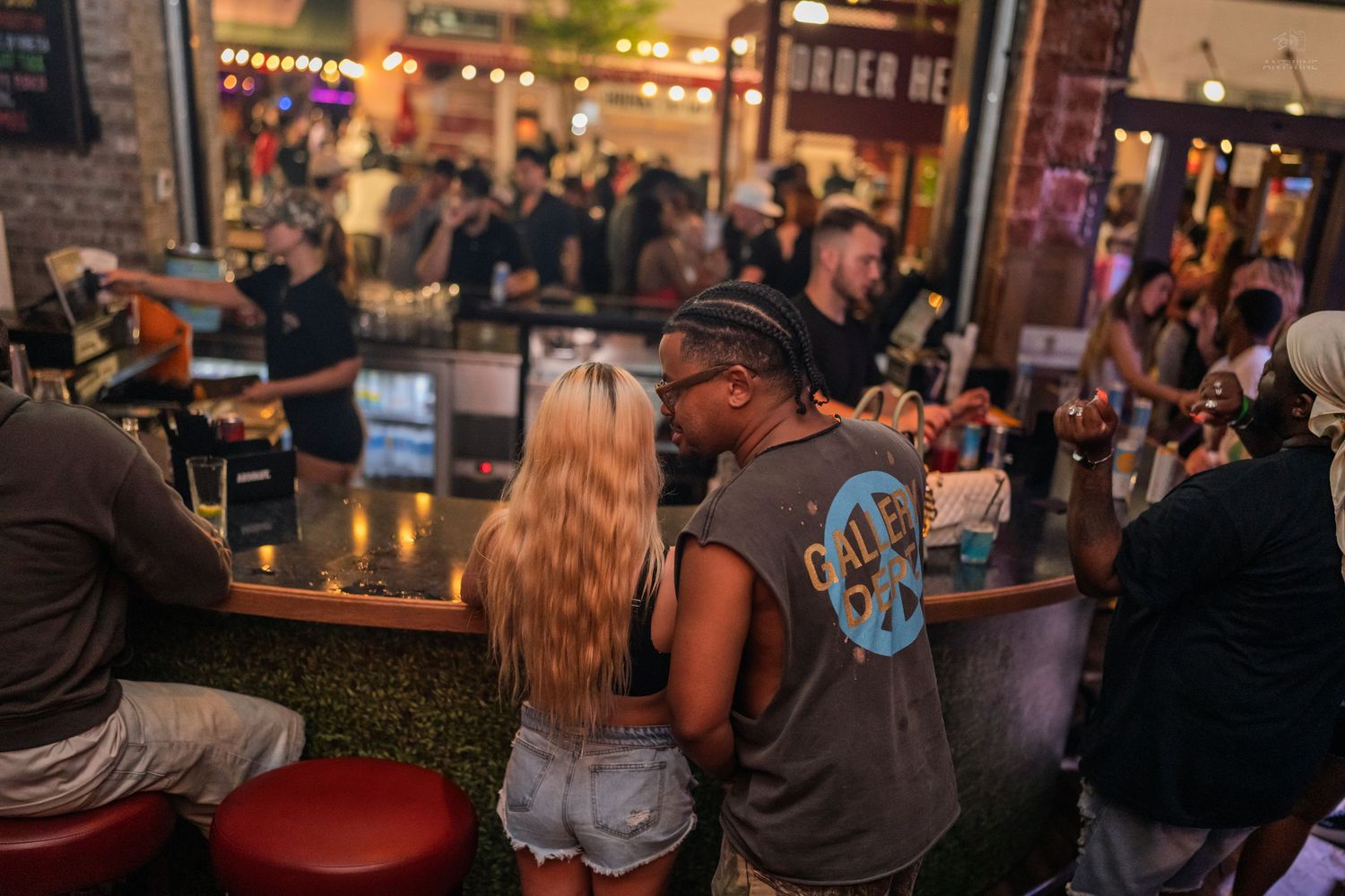 Couple sits closely at an outdoor bar counter at night, surrounded by a lively crowd under string lights.
