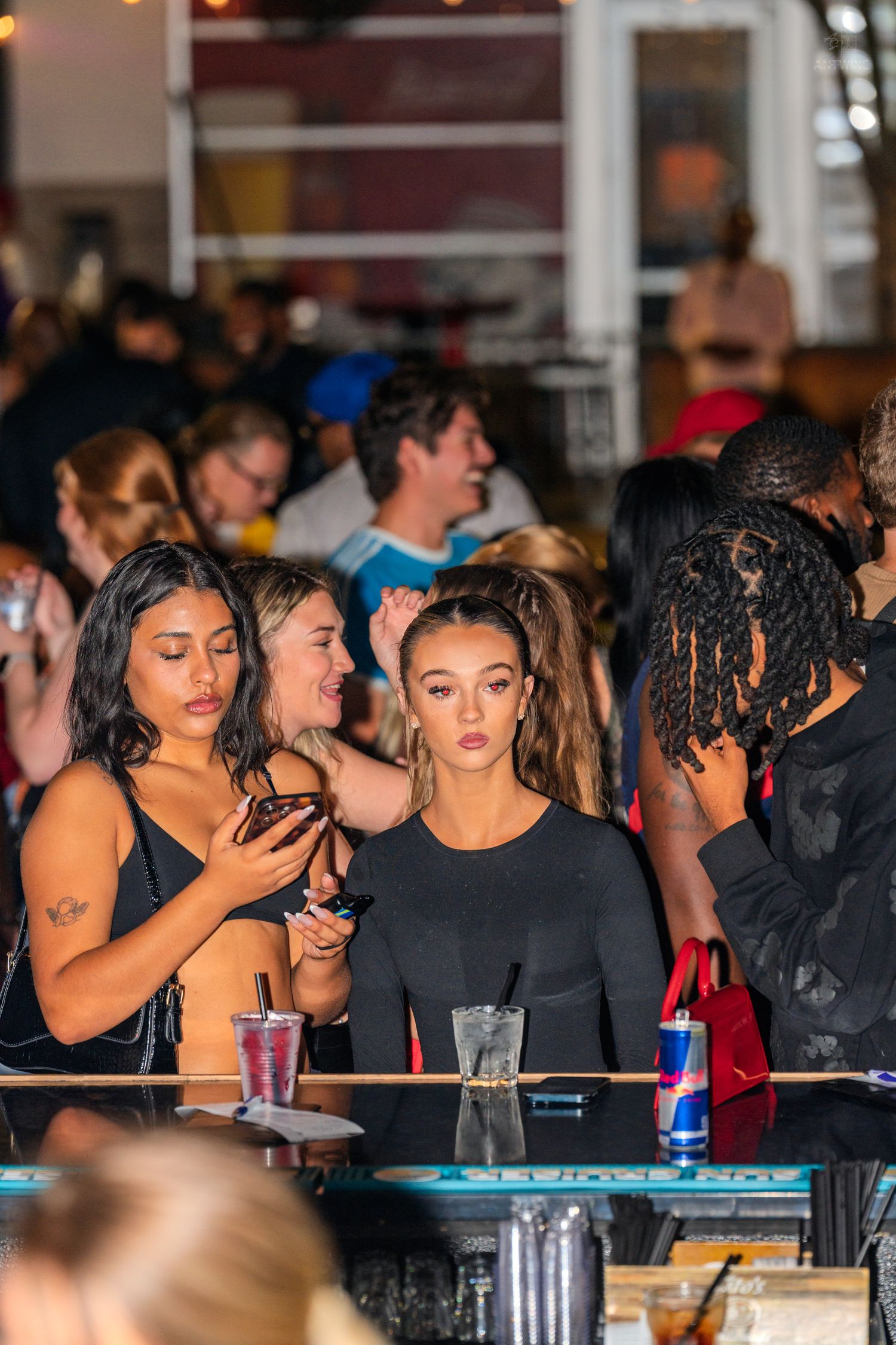 Two women in black outfits standing at crowded bar at nightclub event.